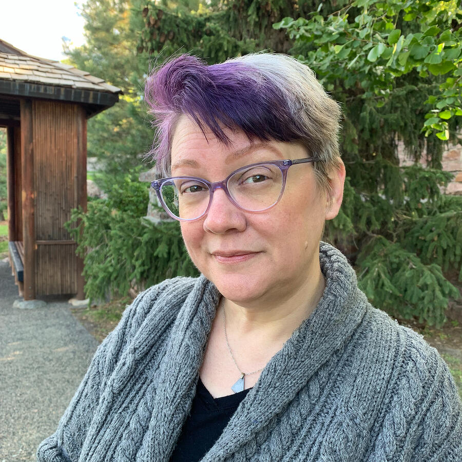 The author: woman with a round face, grey hair, purple bangs, and purple glasses. She's wearing a grey sweater. Trees and a gazebo are behind her.